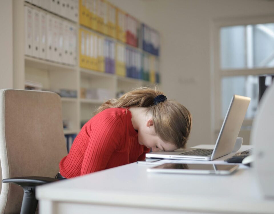 A tired woman in a red sweater leans her head on a desk with a laptop, symbolizing workplace fatigue.