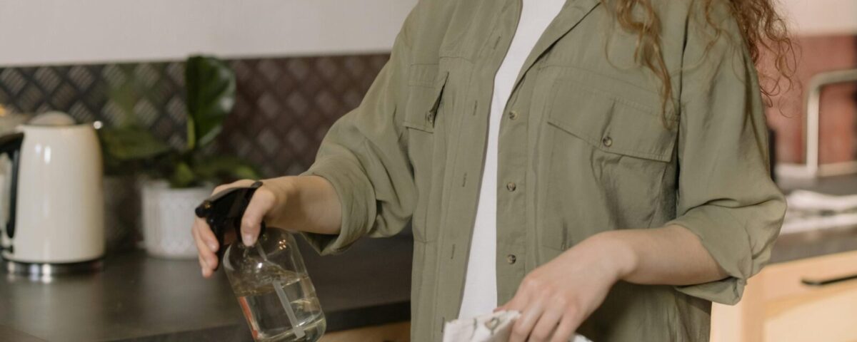 A woman with curly hair cleans a kitchen counter using a spray bottle and cloth.
